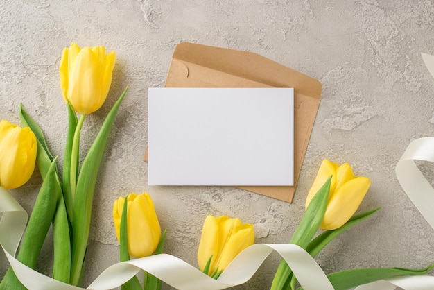 Foto vista dall'alto della busta di carta artigianale della composizione del giorno della donna e mazzo di lettere di tulipani gialli e nastro bianco su sfondo di cemento grigio testurizzato isolato con spazio vuoto