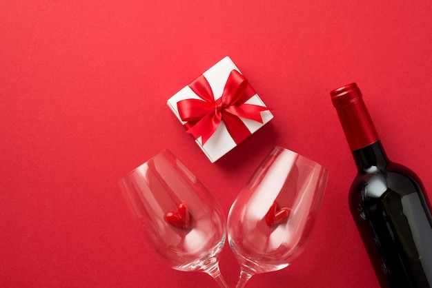 Foto vista dall'alto della confezione regalo con decorazioni di san valentino con fiocco di nastro rosso cuoricini in due bicchieri da vino e bottiglia di vino su sfondo rosso isolato con spazio vuoto