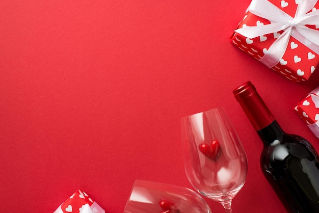 Foto vista dall'alto della confezione regalo di decorazioni di san valentino in carta da regalo rossa con motivo di cuori e piccoli cuori di prua tra due bicchieri di vino bottiglia di vino su sfondo rosso isolato con copyspace