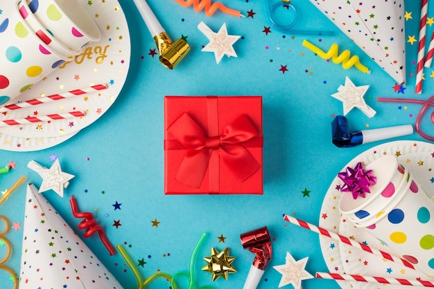 Foto vista dall'alto della confezione regalo rossa con fiocco di nastro al centro della composizione della tavola di compleanno e accessori per feste su sfondo blu isolato