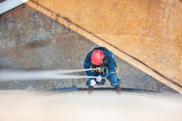 Foto vista dall'alto della saldatrice industriale con accesso alla fune che lavora in altezza indossando l'imbracatura, ispezione dell'accesso alla fune dell'attrezzatura di sicurezza del casco dell'industria dei serbatoi di stoccaggio dello spessore.