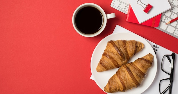 Foto vista dall'alto della tastiera del posto di lavoro raccoglitore rosso clip matita promemoria occhiali tazza di caffè e piatto con croissant freschi su sfondo rosso isolato con spazio vuoto