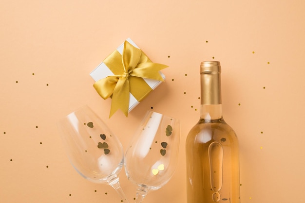 Foto vista dall'alto delle decorazioni di san valentino confezione regalo bianca con fiocco dorato bottiglia di vino due bicchieri da vino con coriandoli a forma di cuore e paillettes su sfondo beige pastello isolato con copyspace