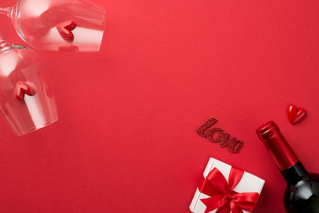 Foto vista dall'alto di decorazioni di san valentino confezione regalo bianca con fiocco rosso cuoricini in due bicchieri da vino iscrizione amore e bottiglia di vino su sfondo rosso isolato con spazio vuoto