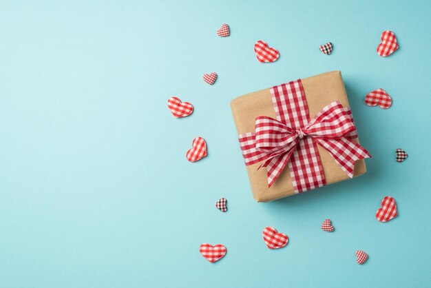 Foto vista dall'alto di decorazioni di san valentino confezione regalo di carta artigianale con fiocco a scacchi circondato da cuori decorativi su sfondo blu pastello isolato con copyspace