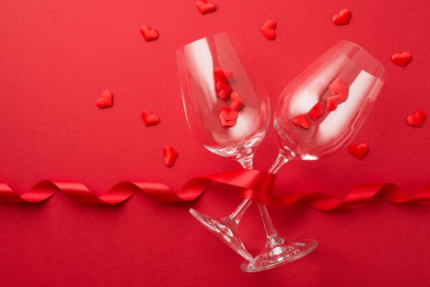 Foto vista dall'alto di decorazioni di san valentino nastro riccio rosso e piccoli cuori che volano da due bicchieri da vino su sfondo rosso isolato con copyspace