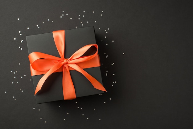 Foto vista dall'alto di elegante confezione regalo nera con fiocco in nastro di raso arancione e paillettes luminose su sfondo nero isolato con copyspace