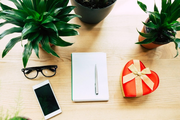 Foto vista dall'alto di smartphone, taccuino bianco, occhiali, piante d'appartamento e confezione regalo su tavola di legno