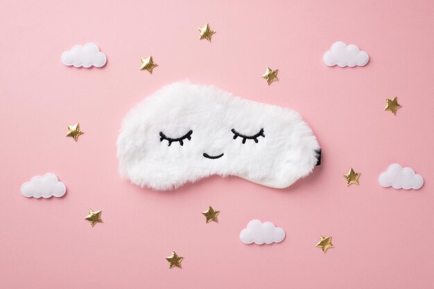 Foto vista dall'alto di una soffice maschera per dormire bianca con occhi chiusi e nuvole sorridenti e stelle dorate su sfondo rosa pastello isolato