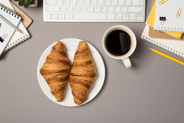 Foto vista dall'alto di workstation tastiera quaderni raccoglitore clip matita gialla penna vaso di fiori tazza di caffè e piatto con due croissant su sfondo grigio isolato