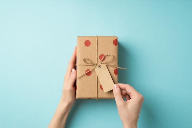 Foto vista dall'alto in prima persona delle mani della ragazza delle decorazioni di san valentino che toccano il cartellino del prezzo su confezione regalo di carta artigianale con motivo a pois e fiocco di spago su sfondo blu pastello isolato con copyspace