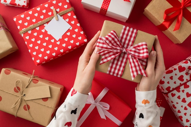 Foto vista dall'alto in prima persona delle mani della ragazza delle decorazioni di san valentino in camicia bianca con scatola regalo di carta kraft con fiocco a scacchi sopra scatole presenti su sfondo rosso isolato