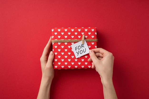 Foto vista dall'alto in prima persona delle mani femminili dell'arredamento di san valentino che prendono nota appuntata sulla confezione regalo in carta da regalo rossa con motivo a cuore e spago su sfondo rosso isolato con spazio vuoto