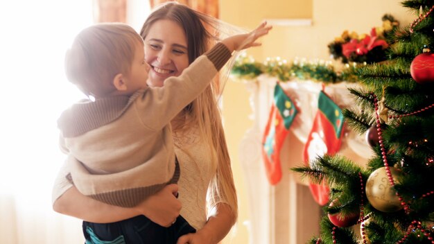 Fotoage 4k di giovane madre felice che tiene e abbraccia il suo piccolo figlio accanto all'albero di Natale in soggiorno. Famiglia che si diverte e si diverte durante le vacanze invernali e le celebrazioni.