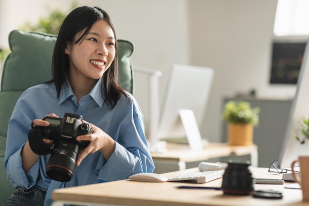 Fotocamera felice della holding del fotografo asiatico della donna che si siede allo scrittorio dell'interno