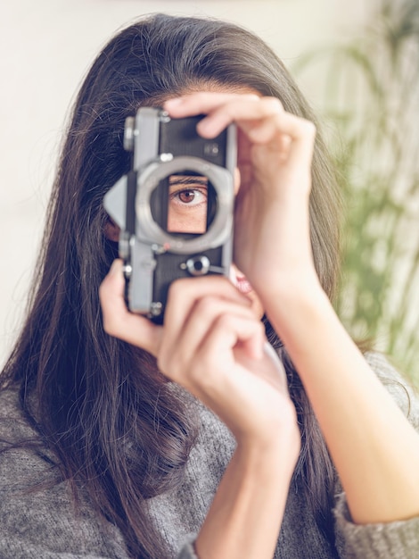 Fotografa donna irriconoscibile con lunghi capelli scuri che guarda attraverso una macchina fotografica a pellicola vintage mentre finge di scattare una foto