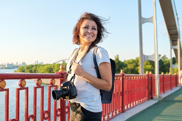 Fotografa matura con fotocamera professionale e zaino, donna sorridente sul ponte in una soleggiata giornata estiva al tramonto. Sfondo di fiume, cielo, skyline della città