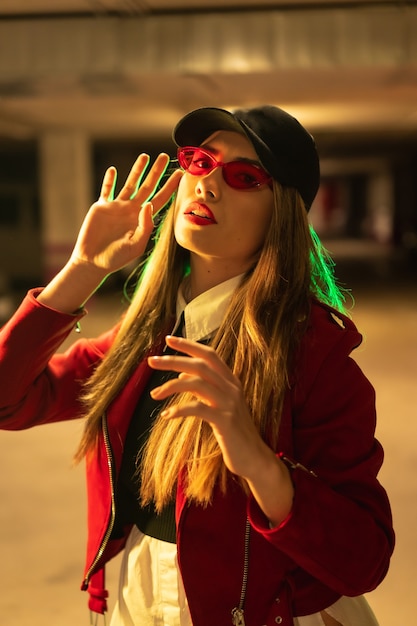 Fotografia con neon rossi e verdi in un parcheggio. Giovane donna caucasica abbastanza bionda in un vestito rosso, occhiali da sole e un berretto nero