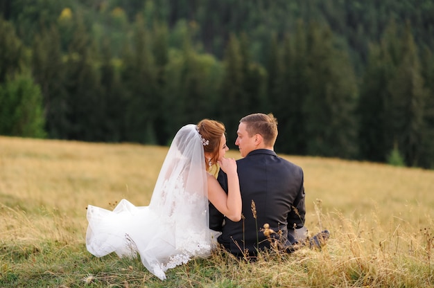 Fotografia di matrimonio in montagna. Sposi seduti con le spalle sull'erba e guardarsi l'un l'altro.