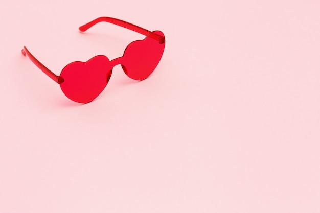 Fotografia di moda stile minimal con cuore a forma di occhiali rossi su sfondo di carta rosa. Occhiali da sole moderni rosa. Concetto estivo alla moda. Copia spazio.