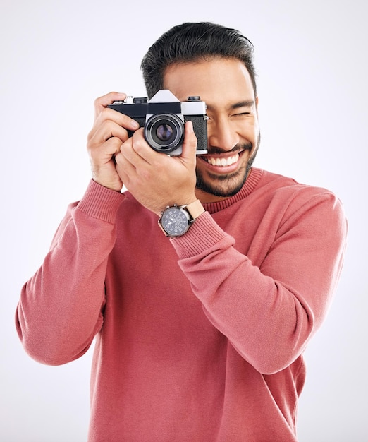 Fotografia felice e uomo con una macchina fotografica in uno studio per la creatività o il lavoro di servizio fotografico artistico Sorriso di felicità e fotografo maschio indiano che scatta foto per paparazzi o media su sfondo bianco