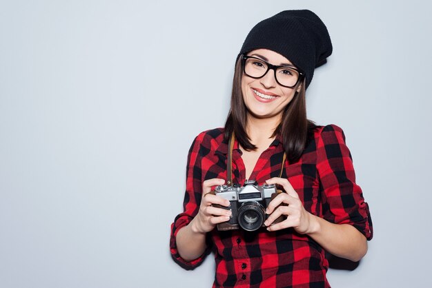 Fotografo alla moda. Bella giovane donna in copricapo e occhiali che tiene la macchina fotografica e guarda la macchina fotografica mentre sta in piedi su uno sfondo grigio