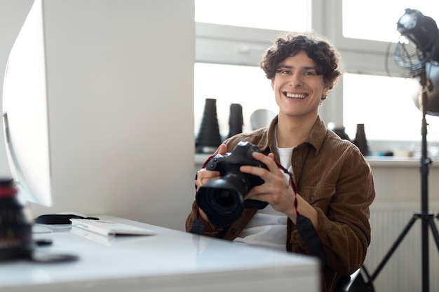 Fotografo allegro del giovane che tiene la macchina fotografica digitale e che sorride alla macchina fotografica che si siede al suo posto di lavoro in studio