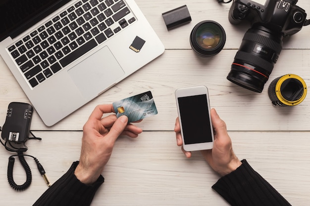 Fotografo che acquista online con carta di credito e smartphone, seduto sul posto di lavoro, ordina nuove attrezzature fotografiche, copia spazio