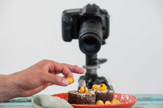 Fotografo che mantiene ciliegia gialla sul dessert, macchina fotografica nella priorità bassa