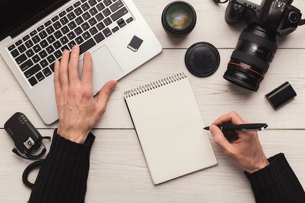 Fotografo che prende appunti durante l'utilizzo del laptop. Vista dall'alto sul posto di lavoro del designer creativo con attrezzatura fotografica e computer, pianificazione degli orari e concetto di autoeducazione, spazio di copia