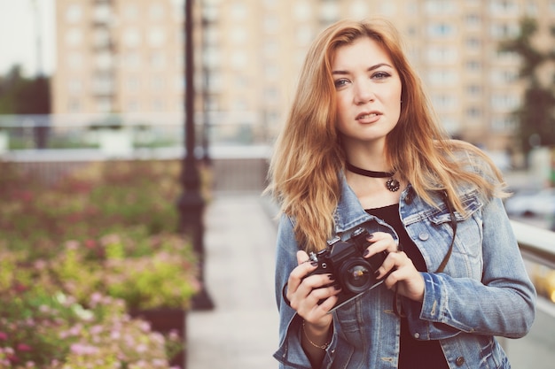 Fotografo di giovane ragazza che cammina lungo la strada in una giacca di jeans con una vecchia macchina fotografica