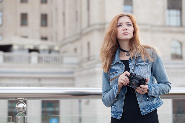 Fotografo di giovane ragazza che cammina lungo la strada in una giacca di jeans con una vecchia macchina fotografica