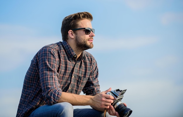 Fotografo hipster. Vlog di tiro per blogger. Bello blogger ragazzo viaggiatore fotocamera retrò. Impostazioni manuali. Blogger di viaggio. Ragazzo all'aperto sullo sfondo del cielo blu. Attrezzatura d'epoca. Giornalista che prende foto.