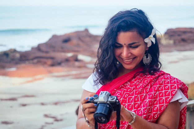 Fotografo indiano femminile che fotografa con la fotocamera digitale sulla spiaggiabella donna indiana in sari tradizionali rossi nel mare paradiso tropicale che scatta foto del servizio fotografico del paesaggio