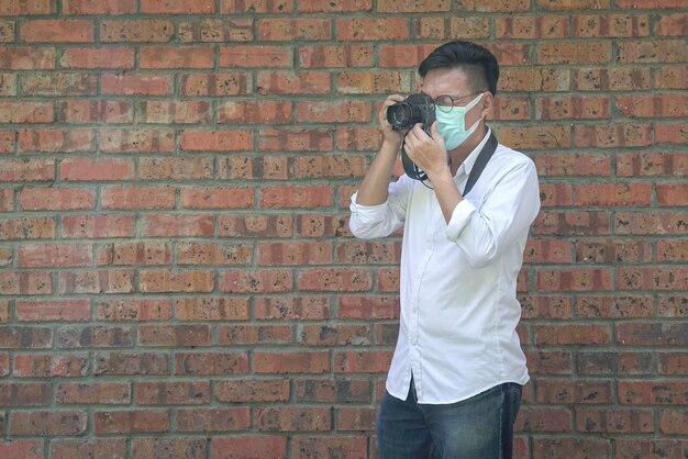 Fotografo maschio che indossa una maschera facciale in camicia bianca e jeans Sfondo muro di mattoni