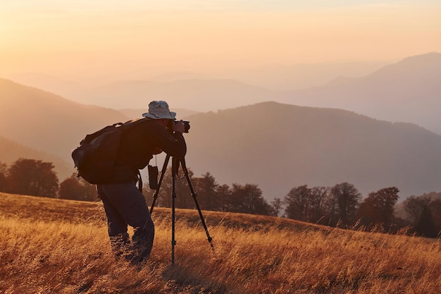 Fotografo maschio in piedi e che lavora al maestoso paesaggio di alberi autunnali e montagne all'orizzonte