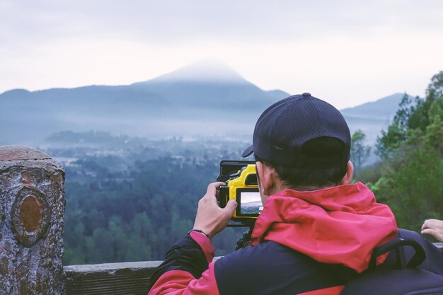 fotografo paesaggista che lavora con fotocamera reflex digitale e treppiede indossando giacca cappello e zaino in cima