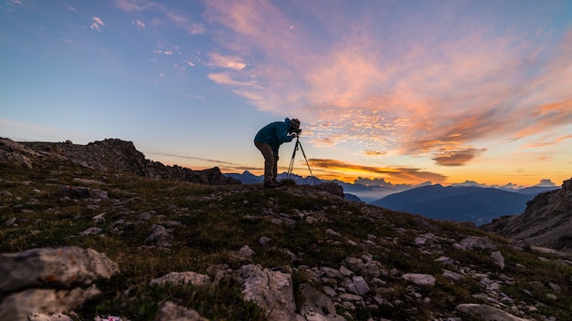 Fotografo sulla cima della montagna con la macchina fotografica sul treppiede al paesaggio variopinto leggero di scenis del cielo di alba.