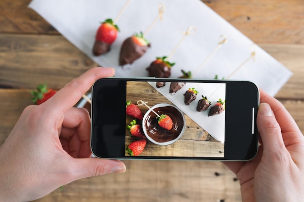 Fragole con cioccolato su uno sfondo di legno Mano che scatta foto con il cellulare