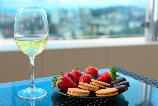 Fragole e dolci sul piatto con un bicchiere di champagne o vino bianco su un tavolo sul balcone o sulla terrazza con vista sfocata sui tetti della città e sul mare Messa a fuoco selettiva Concetto di viaggio di stile di vita di lusso