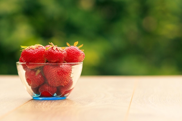 Fragole mature fresche in ciotola di vetro su tavola di legno con bokeh naturale verde sullo sfondo