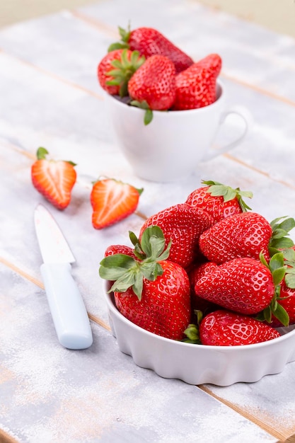 Fragole succose in una ciotola di ceramica bianca e frutti di bosco a fette su un tavolo di legno grigio chiaro