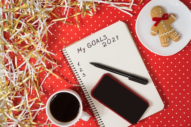 Frase i miei obiettivi 2021 in un taccuino, penna nera e smart phone. Pan di zenzero e caffè su sfondo rosso.