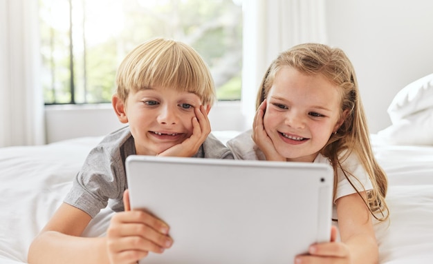Fratelli e sorelle felici si rilassano e guardano con il tablet a letto per spettacoli di intrattenimento online o cartoni animati a casa I bambini piccoli fratello e sorella sdraiati con la tecnologia per serie di film o media in camera da letto
