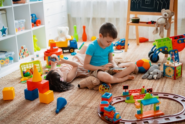 Fratelli e sorelle, fratelli e sorelle, gli amici si siedono nella sala giochi per bambini della casa al piano terra con gli smartphone, staccati dai giocattoli sparsi.