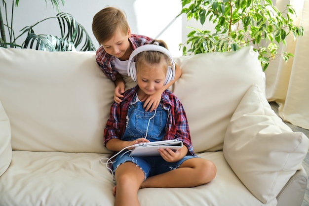 Fratello e sorella carini con le cuffie che si godono il tablet a casa. famiglia, bambini, tecnologia e concetto di casa