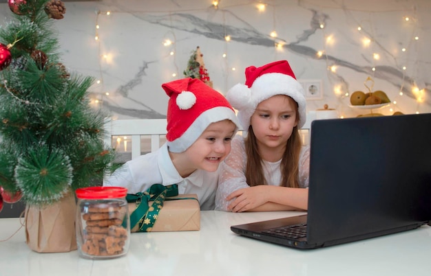 Fratello e sorella con il cappello di Babbo Natale si divertono. Bambini a tavola con laptop e regali, chiamano amici o parenti tramite collegamento video e salutano. La famiglia sta festeggiando il Natale a casa. Riduzione dei contatti.