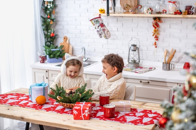 Fratello e sorella dei bambini in cucina che celebrano il Natale. bambini felici al tavolo della cucina aprono doni. momenti meravigliosi di apertura di un regalo a sorpresa