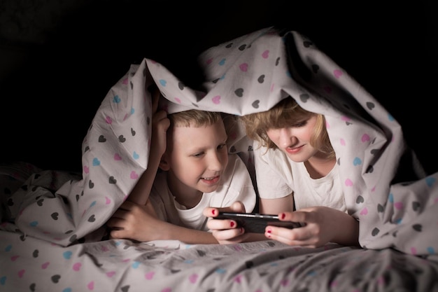 Fratello e sorella si sdraiano sotto una coperta di notte e giocano al telefono