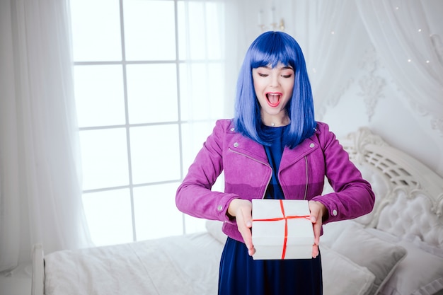 Freak alla moda. Il glamour ha sorpreso che la bella donna con capelli blu sta tenendo il regalo nella camera da letto bianca. Concetto di moda e bellezza